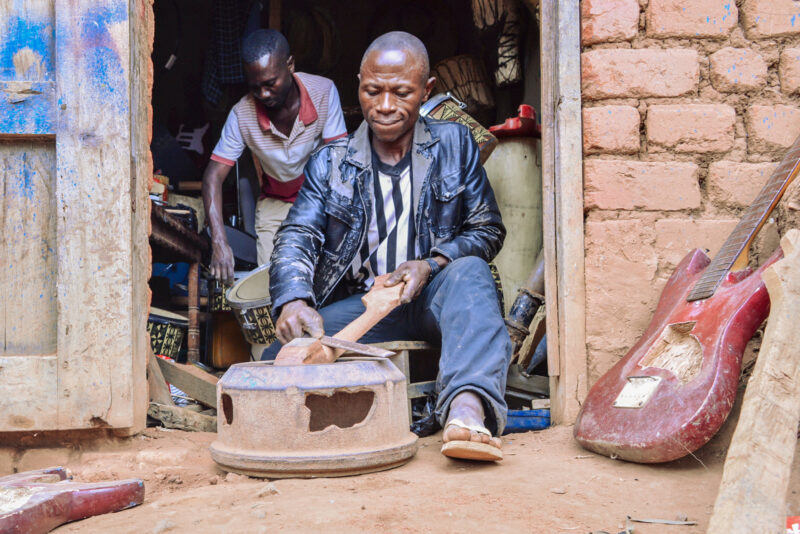 Moise Muhindo Kisuba construit une guitare dans son atelier de Kirumba, au sud du territoire de Lubero en République démocratique du Congo.  Derrière lui, Egide Kasereka Kighoma, un apprenti, travaille sur un tambour le 15 juillet 2021. Crédit : Zita Amwanga/Global Press Journal.