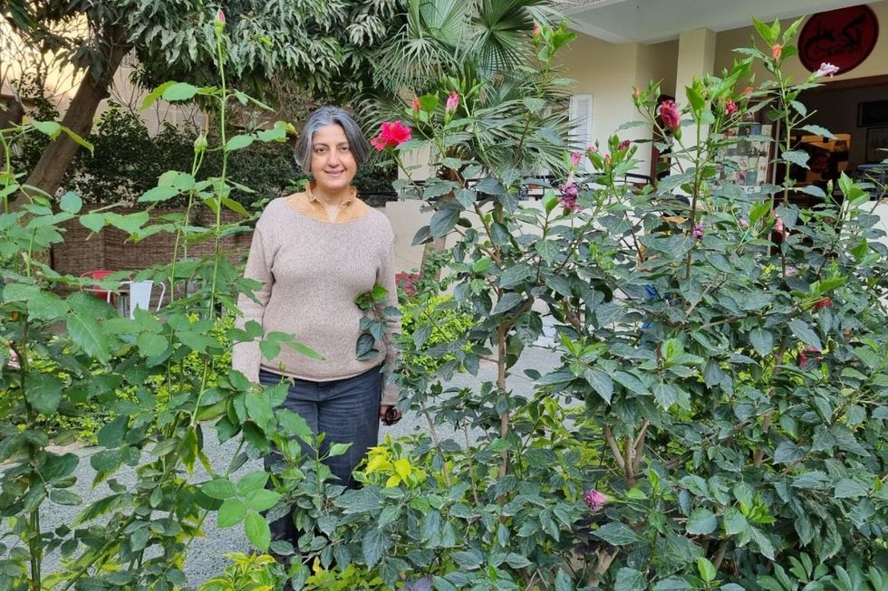 Karam Youssef se tient devant l'entrée de sa librairie, derrière un parterre de fleurs avec des plantes hautes et des fleurs rouges. Elle porte un pull gris clair et un pantalon gris foncé.