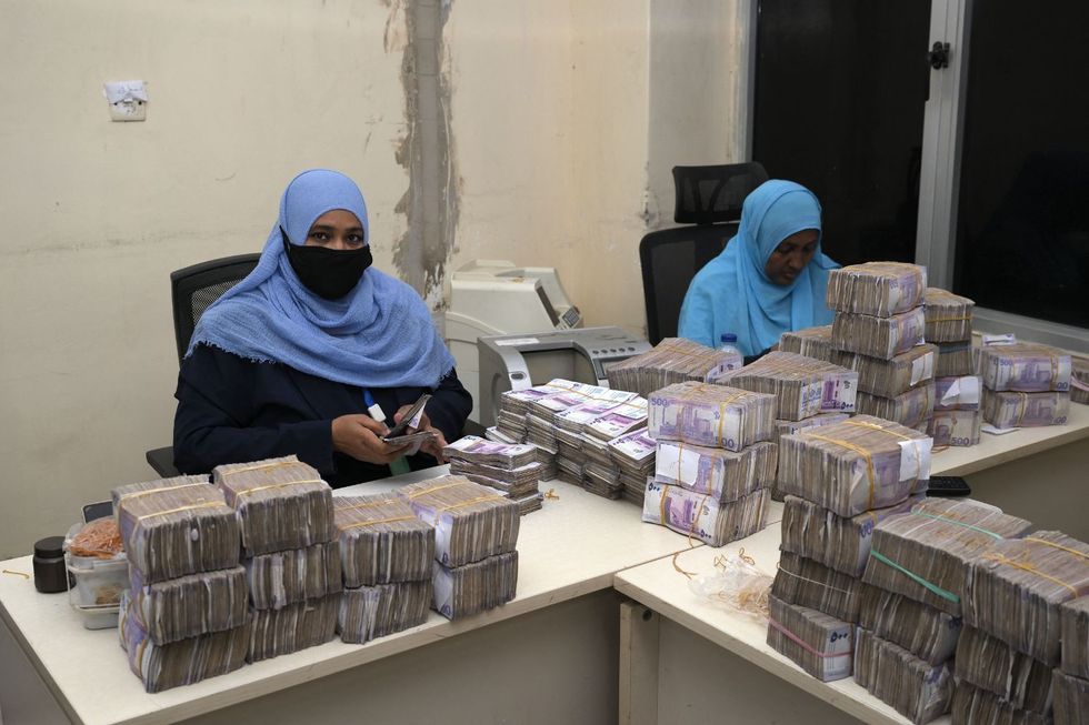 Deux femmes portant des hijabs bleus sont assis derrière des bureaux blancs, comptant de grandes piles de factures