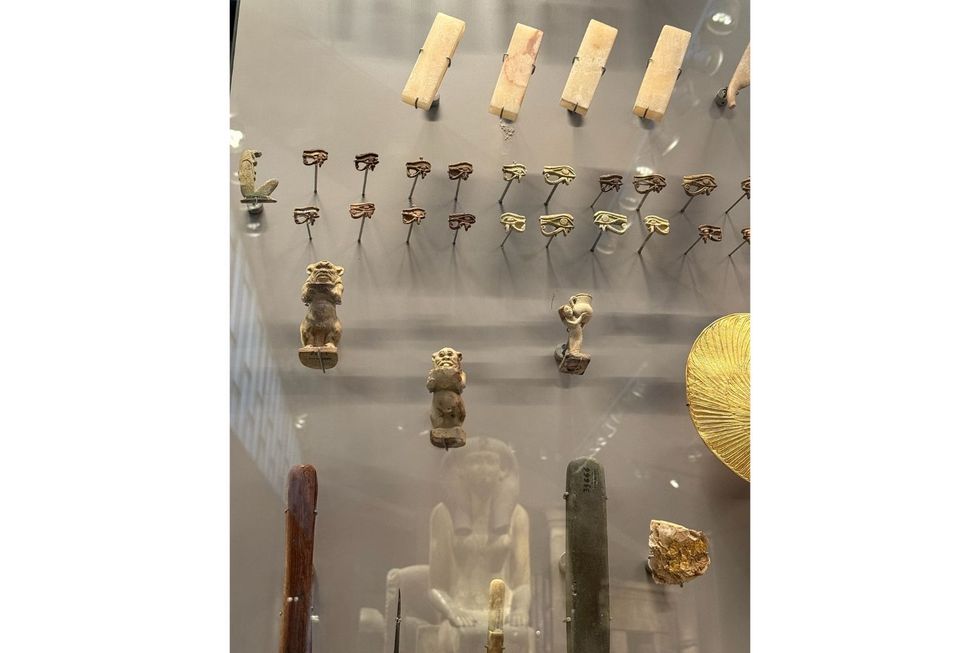 Une vitrine avec des artefacts anciens, comme l'œil d'Horus et un petit singe, et le reflet d'une statue ancienne.