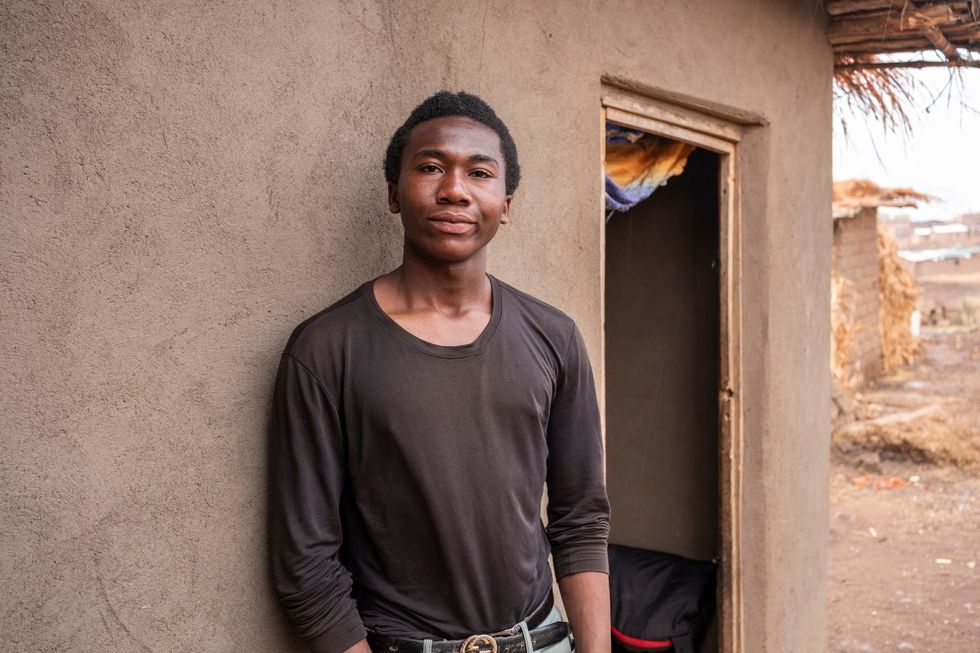 Espoir Kahitani, un jeune homme en chemise noire, se tient à côté d'une maison dans le camp de réfugiés de Dzaleka.