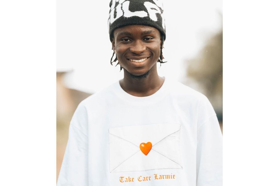 Datway Ezzy est vêtu d'un t-shirt blanc avec les mots  u201take care larmie  u201d et un chapeau noir et blanc, il sourit et regarde directement la caméra.