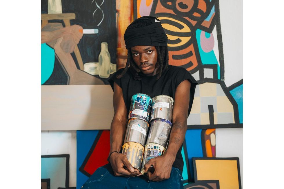 Gerald Massaquoi se trouve devant une murale colorée, tenant des canettes de peinture. Il porte un bonnet noir, un t-shirt noir et un jean bleu.