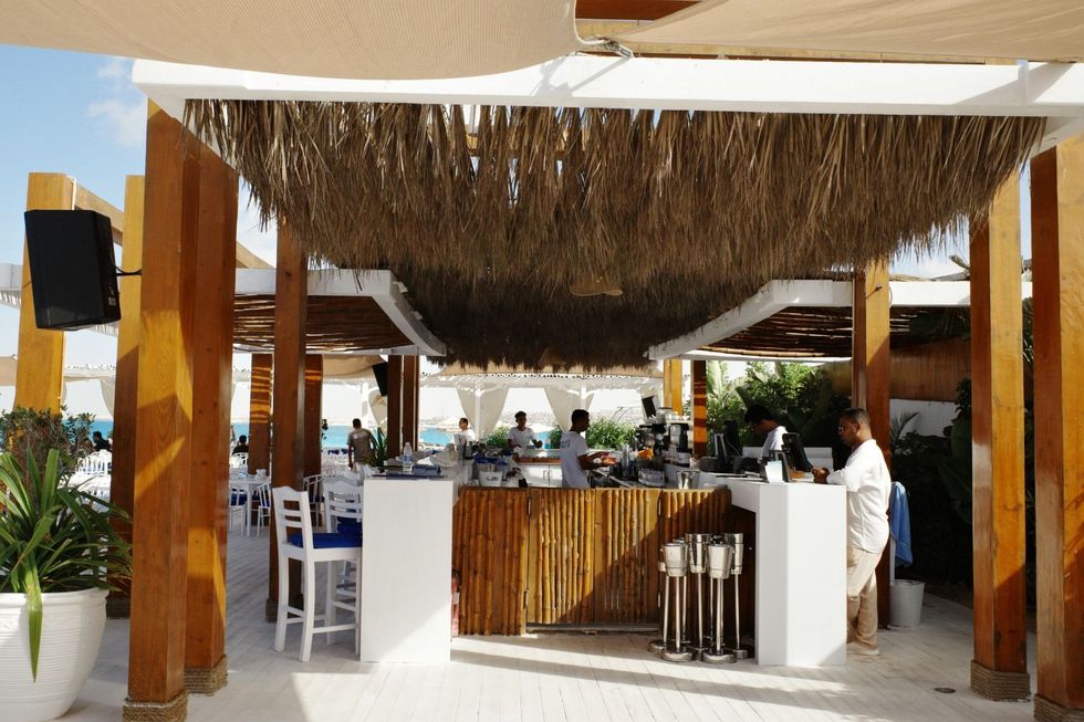 Le bar d'un restaurant de plage, en bois, des pailles en bambou et des textiles blancs. La Méditerranée bleue est en arrière-plan.