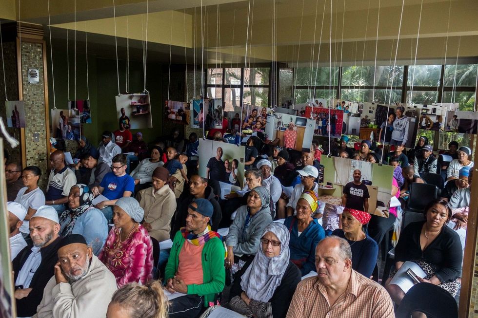 Des hommes et des femmes de couleur et de foi différentes s'asseyent dans une salle avec des photographies suspendues au-dessus de leur tête