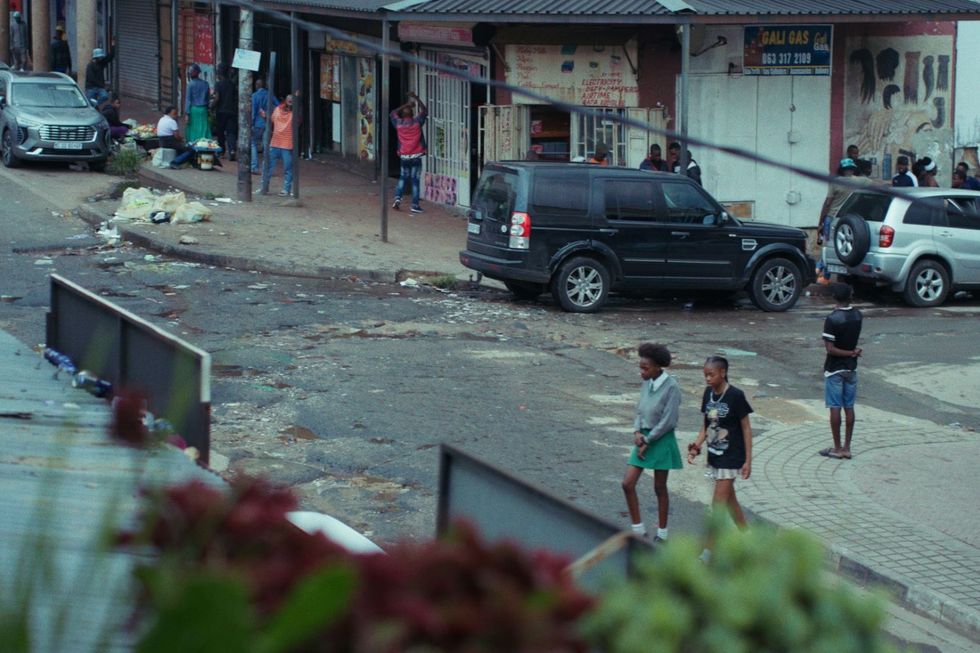 Une vue sur la rue Rocky de Yeville  U2019 à partir de la fenêtre de Sandile, montrant des gens dispersés et quelques véhicules garés sur le bord de la route.