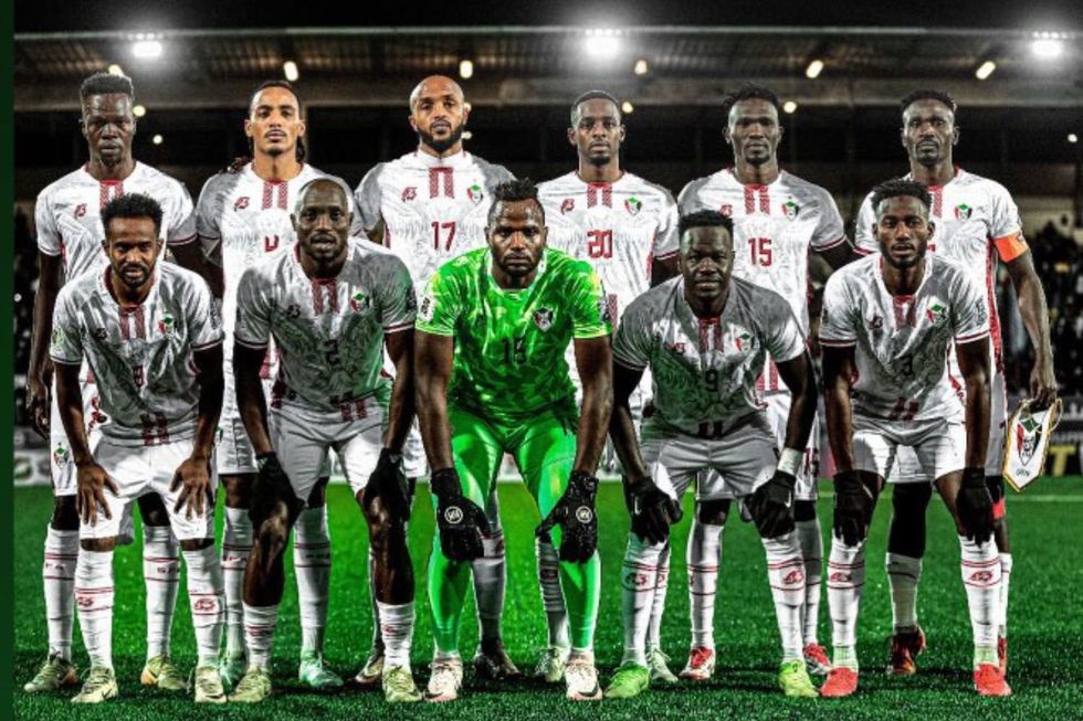 L'équipe nationale de football du Soudan posant pour une photo de groupe sur le terrain avant un match. Les joueurs sont vêtus de maillots blancs avec des détails rouges, à l'exception du gardien de but qui porte un kit vert vif. L'équipe est disposée en deux rangées, avec la première rangée agenouillée et la rangée arrière debout.