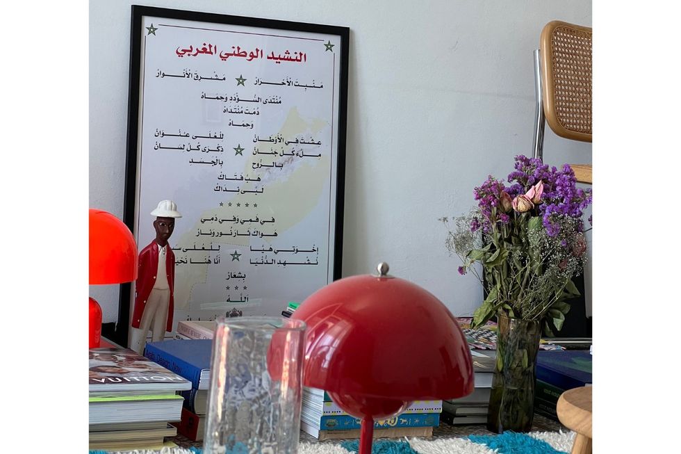 Une affiche avec une police arabe et une statue d'un homme noir sont sur le sol par un mur, aux côtés de livres, de lampes rouges et d'un bouquet de fleurs.