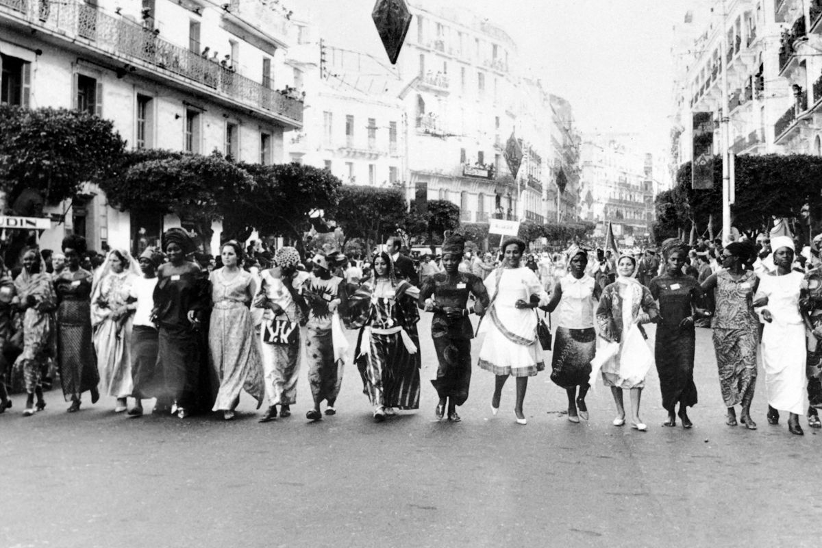 Photo en noir et blanc d'un groupe de femmes dans des tenues africaines et occidentales traditionnelles bras de marche dans un bras dans une rue en Algérie, avec des foules qui regardent des bâtiments voisins.