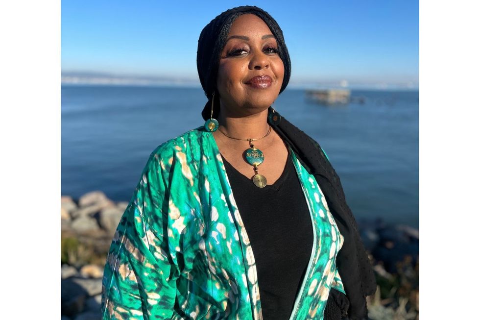 Le journaliste de radio américain soudanais Hana Baba se tient devant la mer, portant un kimono vert, des bijoux verts assortis et un hijab noir. Elle sourit dans la caméra.