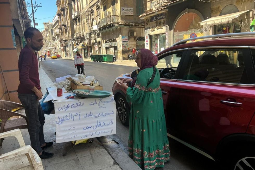 Samah Saeed et son neveu Mohamed Salah vendent Feseekh dans une rue Alexandrie. Elle porte une robe traditionnelle verte avec un hijab rouge et compte de l'argent car elle sert un client dans une voiture rouge.