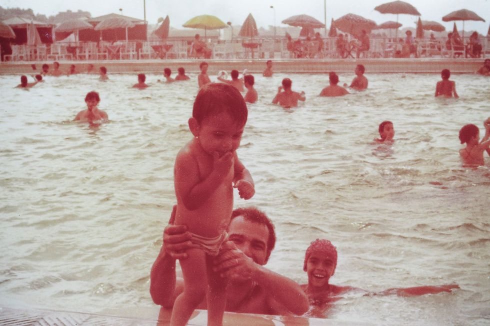 Vieille image sépia d'un homme dans une piscine publique, tenant un tout-petit qui se tient au bord de la piscine. Derrière lui se trouve une jeune fille qui rit.