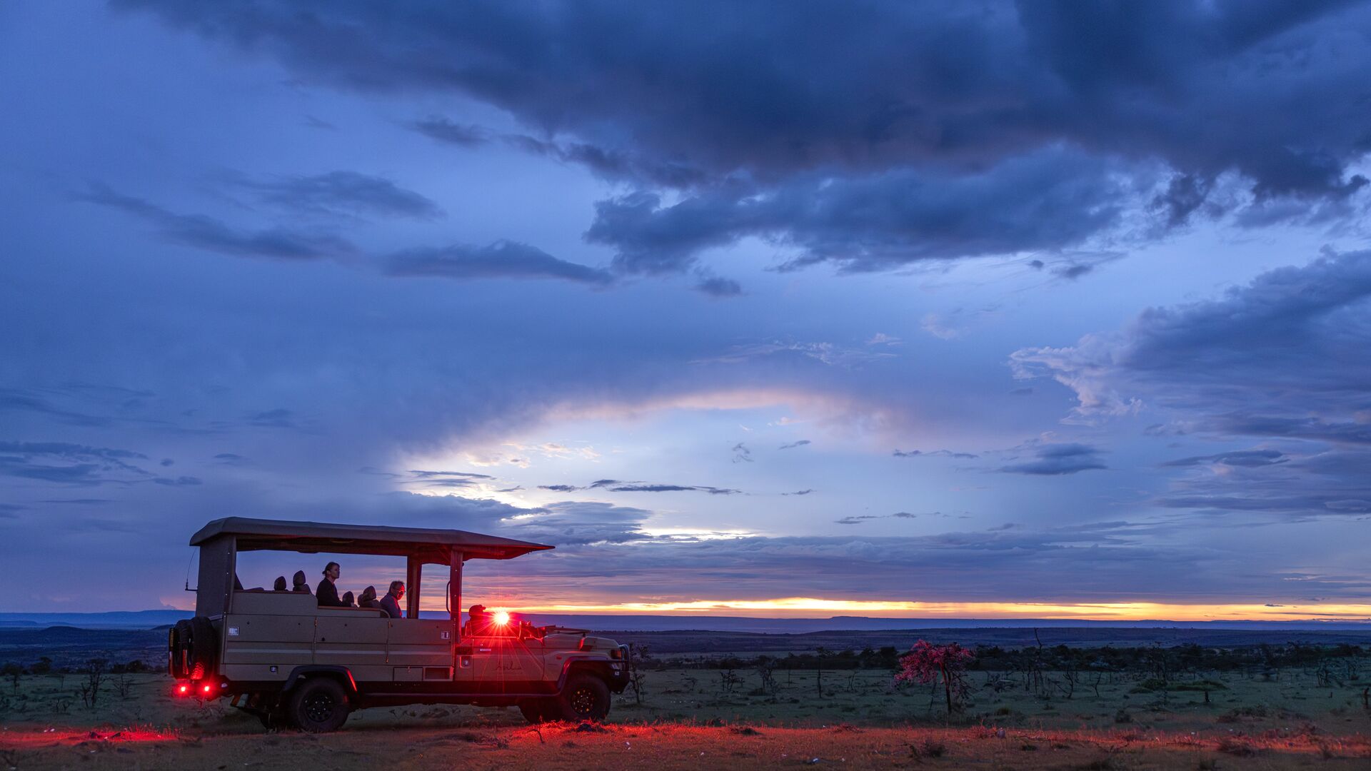 Les invités montent à bord d'un véhicule de safari ouvert lors d'un safari nocturne depuis le camp Naboisho, éclairé par des lumières rouges sur une vaste savane et un ciel crépusculaire spectaculaire.