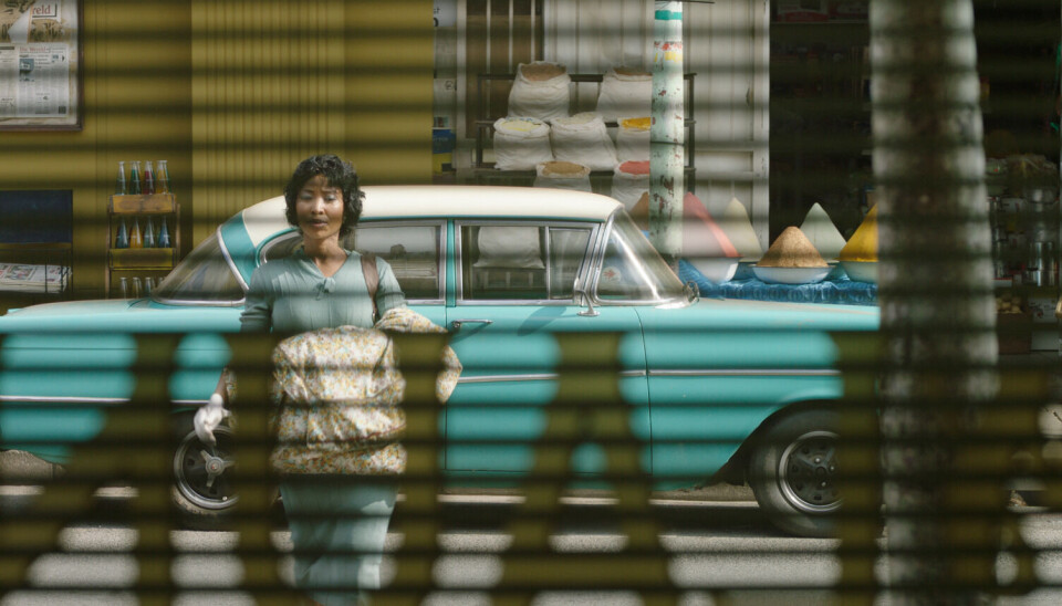 Une image promotionnelle du film Laundry montrant l'un des acteurs vu à travers des stores. Une Chevrolet couleur sarcelle avec un toit blanc est garée derrière elle.