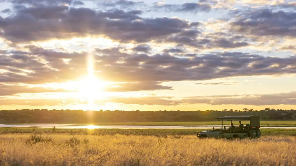 Oliver's Camp, Tarangire, un véhicule de safari ouvert transportant des invités s'arrête dans de hautes herbes dorées au coucher du soleil, surplombant une voie navigable calme avec des nuages ​​spectaculaires et une lumière chaude du ciel