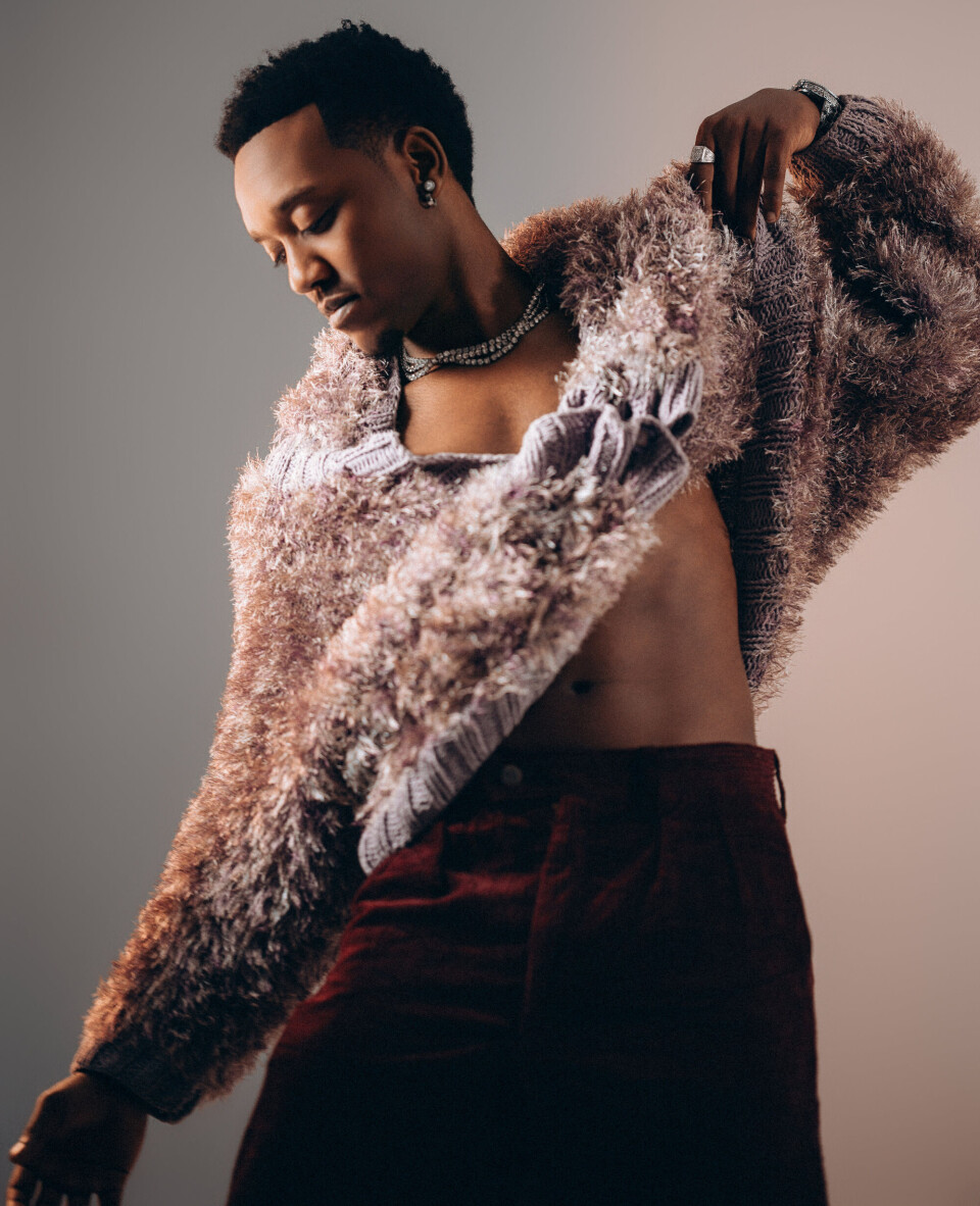 L'artiste et producteur rwandais Element Eleéeh pose artistiquement dans un pull texturé et un pantalon bordeaux.