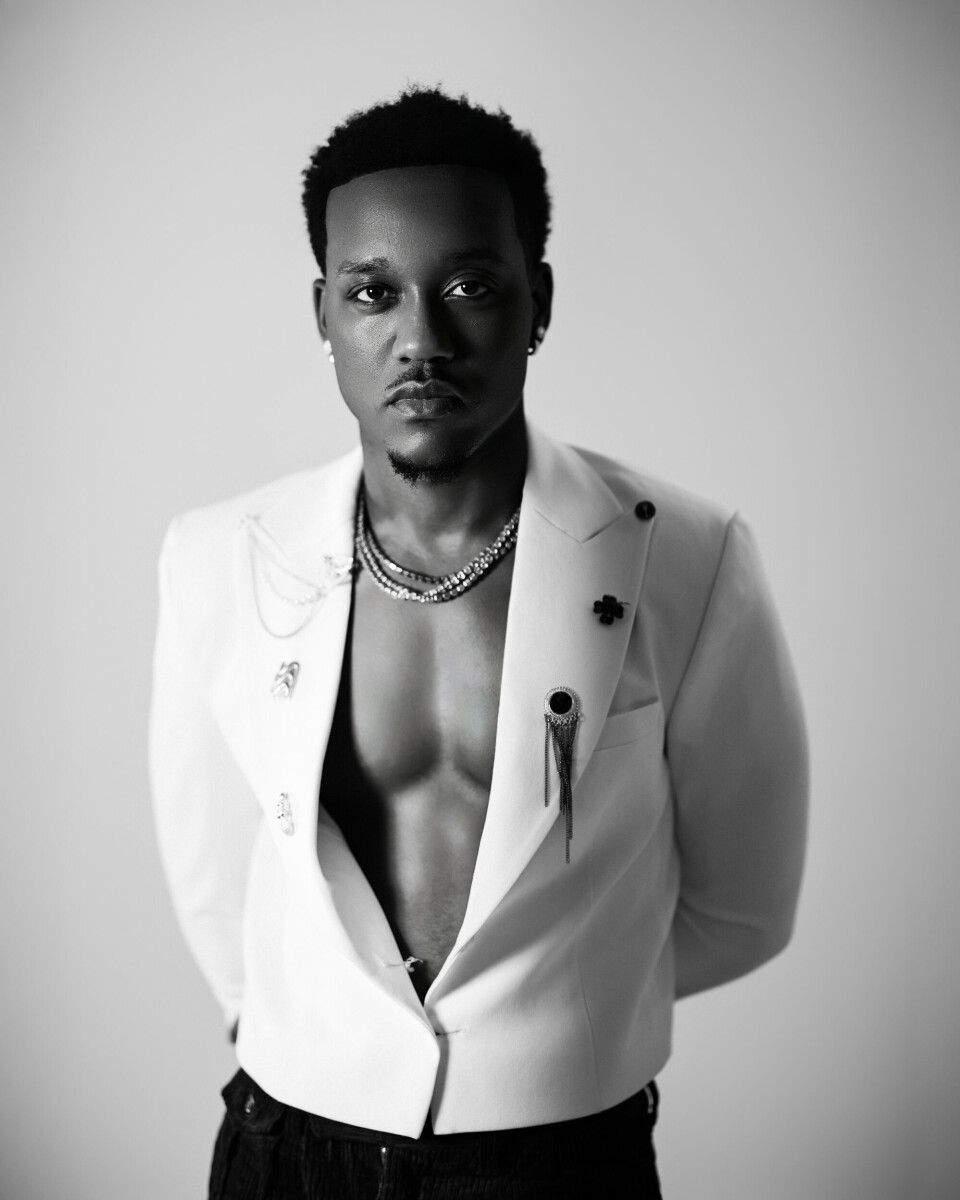 Portrait en noir et blanc de l'artiste et producteur rwandais Element Eleéeh portant un blazer blanc avec la poitrine apparente.