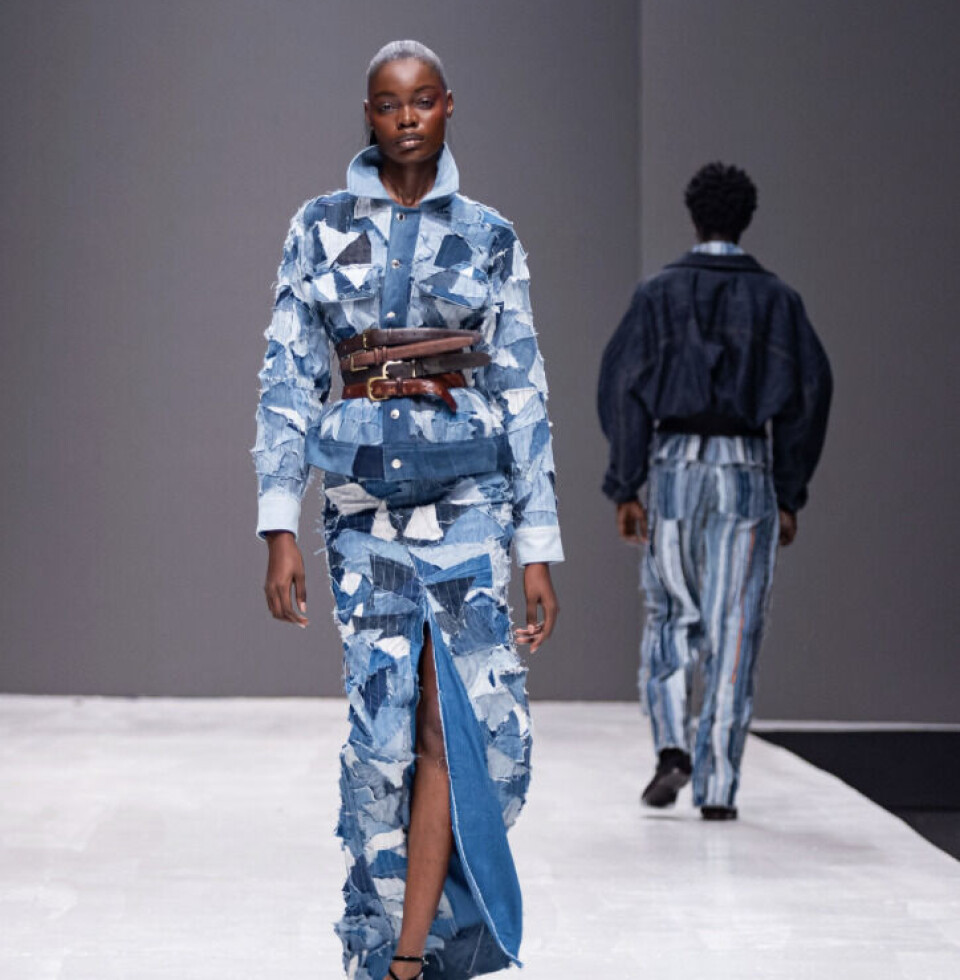 Les créations Boyedoe présentées sur le podium de la Fashion Week de Lagos. Un mannequin sur le podium portant Boyedoe. Sa tenue est une robe-manteau en denim patchwork bleu avec une ceinture en cuir marron.