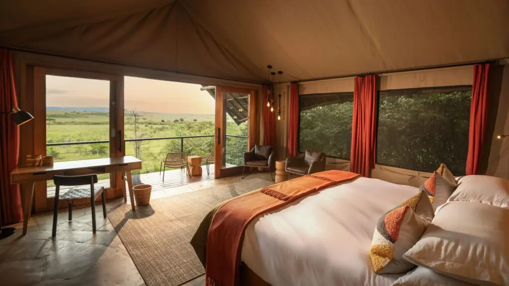 Intérieur d'une chambre double safari de luxe au camp Naboisho avec un lit king-size, un mobilier chaleureux, de grandes fenêtres et une terrasse privée donnant sur la savane ouverte.