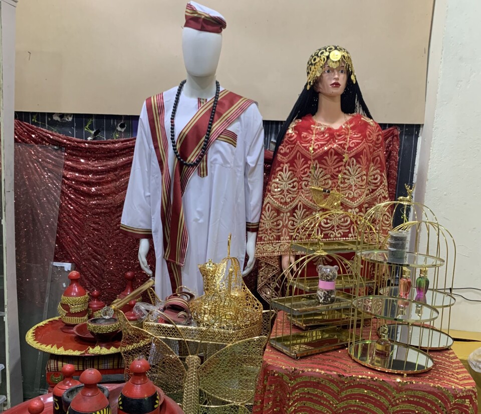 Tout l’attirail nécessaire est disponible au Caire, où des femmes récemment déplacées ont mis à profit leurs compétences entrepreneuriales et bâti une économie du mariage. Deux mannequins vêtus d'une tenue jirtig traditionnelle et debout derrière des tables et des plateaux dorés et rouges portant l'attirail du jirtig.