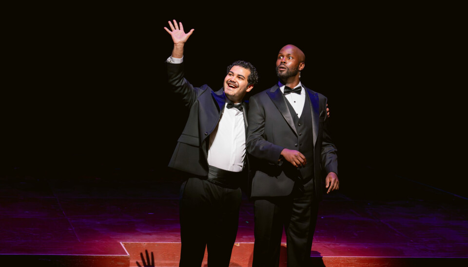 Deux artistes d'opéra en smoking noir se tiennent sous les lumières de la scène ; l'un sourit et salue tandis que l'autre regarde vers l'extérieur, un bras autour de son collègue.