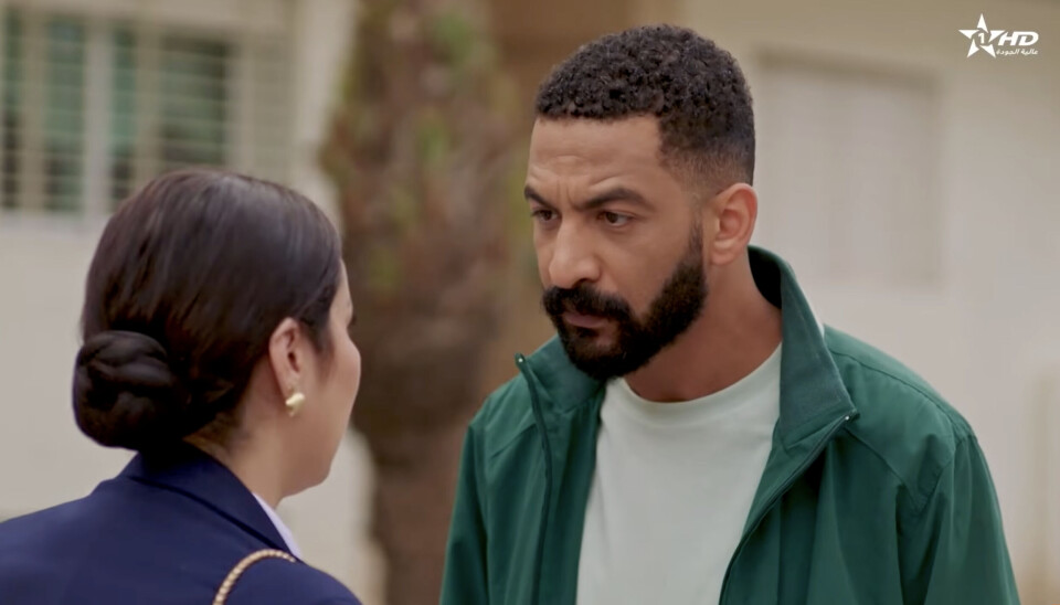 Un homme vêtu d’une veste verte et d’une chemise grise parle à une femme qui porte ses cheveux en chignon soigné. Son expression faciale est sérieuse et agacée.