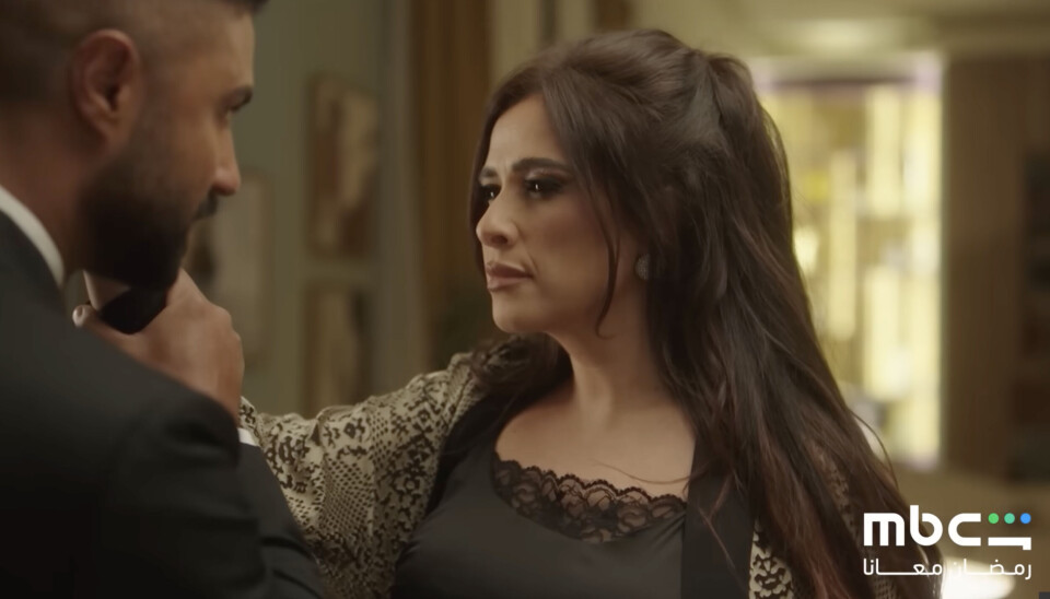 Une femme aux longs cheveux bruns est sur le point de frapper un homme en costume au visage, mais celui-ci lui arrête le bras et lui tient la main juste à côté de sa joue.