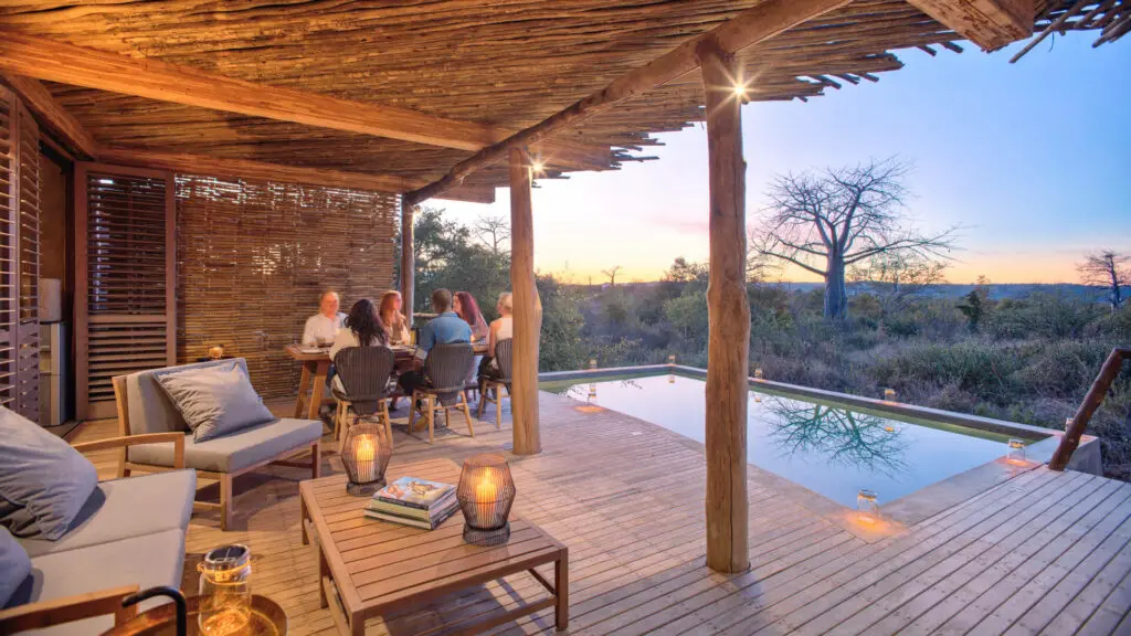 Maison privée Jabali, parc national de Ruaha, invités en train de dîner au bord de la piscine éclairée par des lanternes au coucher du soleil, chaises longues, table avec livres et bougies