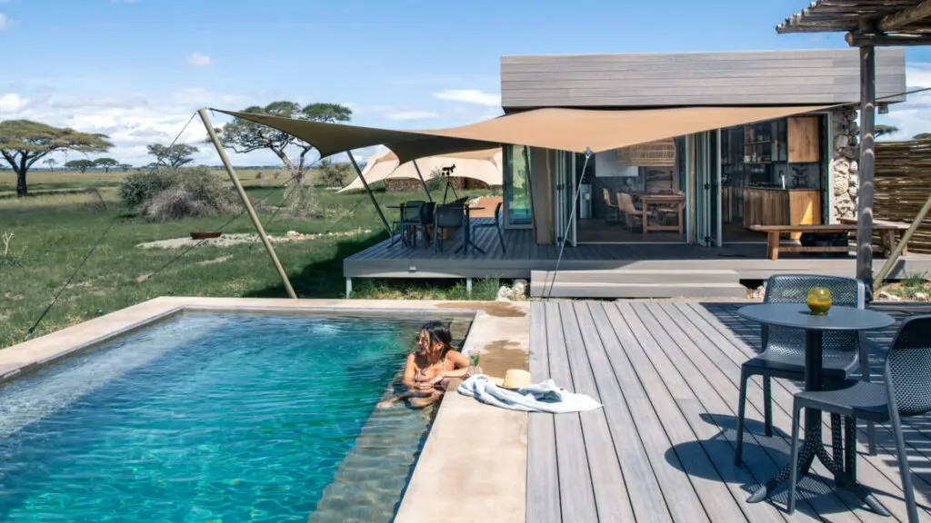 Namiri Plains Retreat, parc national du Serengeti, invité dégustant un cocktail dans la piscine, zone principale en arrière-plan, table et chaise avec bougie en verre shanga sur la table sur la terrasse