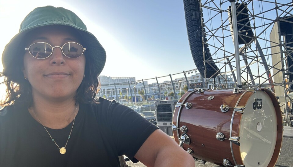 Reda prend un selfie avec un drôle de sourire, assis sur une scène extérieure, portant un chapeau vert et des lunettes de soleil. A côté d'elle se trouve un grand tambour.