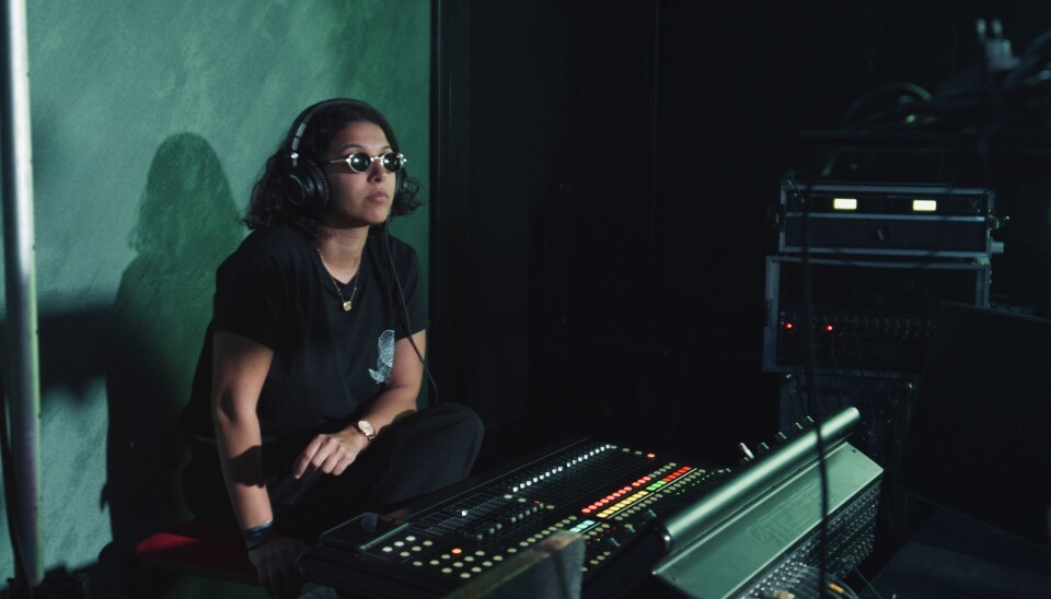 Reda porte une chemise noire, des lunettes de soleil et de gros écouteurs noirs, assis derrière un grand contrôleur avec une expression concentrée et sérieuse.