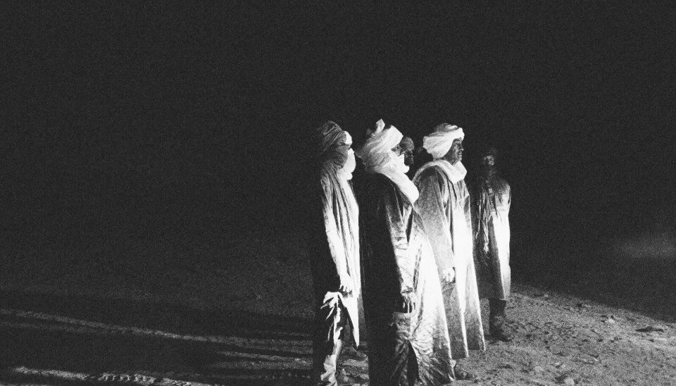 Cinq hommes en tenue traditionnelle touarègue se tiennent la nuit dans le désert. Une lumière vive les éclaire de face et projette les ombres de leurs personnages derrière eux lorsqu'ils la regardent.