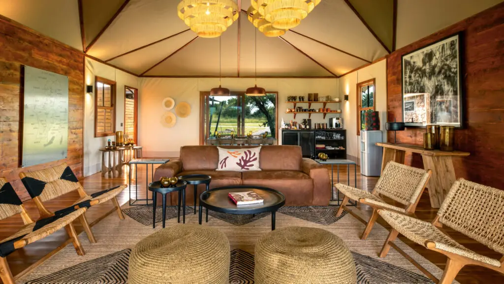 Sayari Retreat, parc national du Serengeti, espace principal partagé avec canapé, chaises en osier, tables, livres, ornements en verre Shanga, bar et cuisine à l'arrière