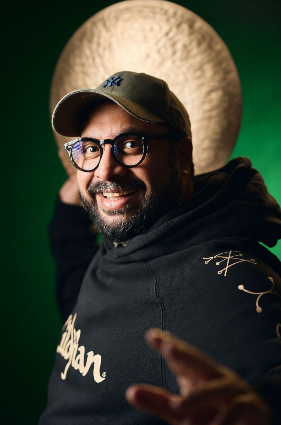 Une image de profil de Kesivan Naidoo regardant la caméra tout en tenant une cymbale derrière la tête. Il porte une casquette NY olive et un pull azerbaïdjanais noir.