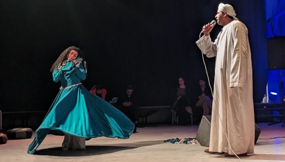 Une femme virevolte dans une longue robe verte tandis qu'un homme vêtu d'une jalabiya blanche et d'un turban chante à côté d'elle.