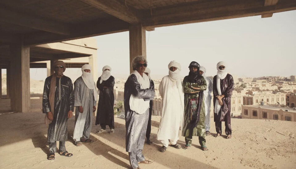 Neuf hommes en tenue traditionnelle touarègue, dont huit portent des turbans, se tiennent sous un bâtiment surélevé, avec des maisons plus traditionnelles en arrière-plan.