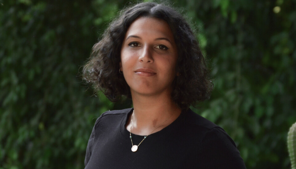 Portrait de Reda portant un t-shirt noir, un collier doré et de petites boucles d'oreilles dorées, souriant à la caméra devant un fond vert luxuriant.