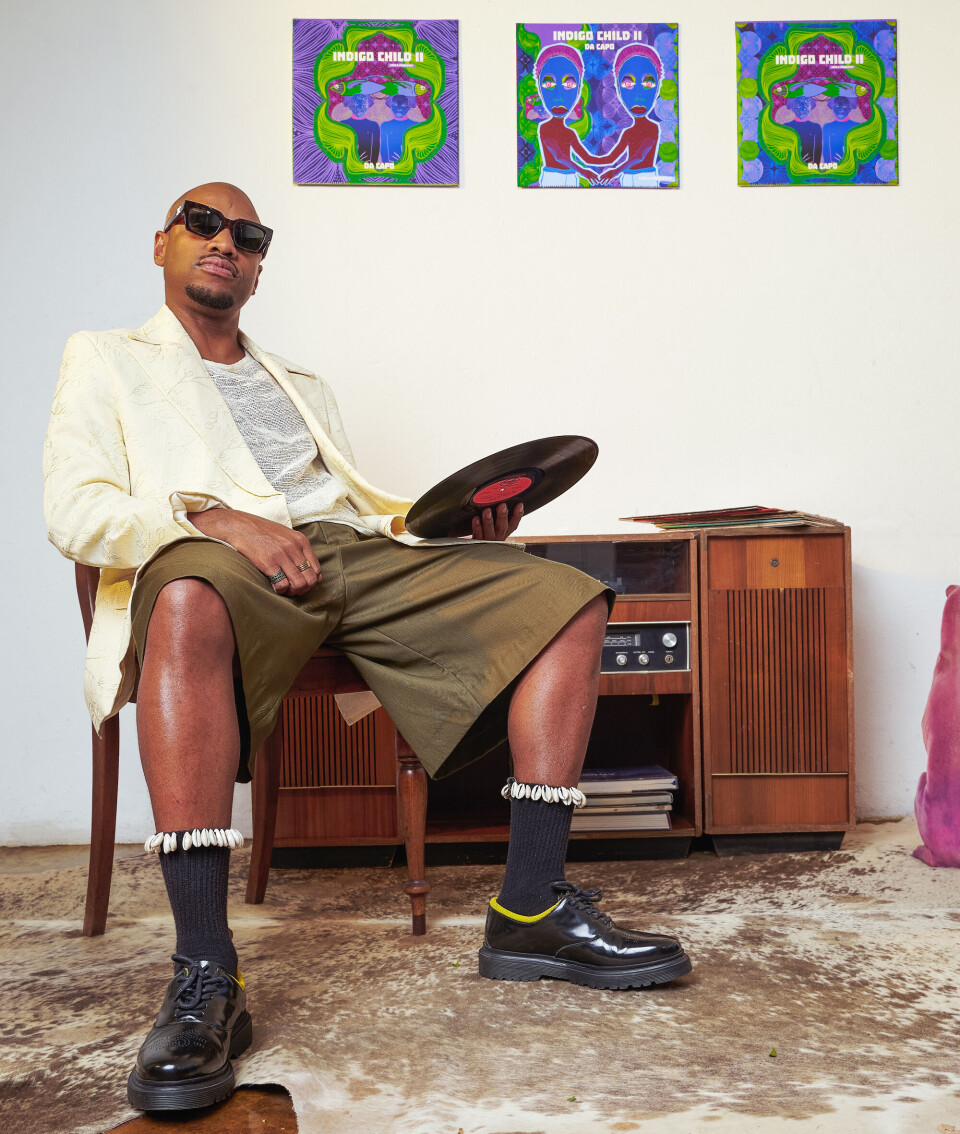 Da Capo tient un disque vinyle alors qu'il est assis dans une image de presse. Derrière lui se trouve l'illustration d'Indigo Child II et il porte un blazer blanc crème, un short olive et des chaussures noires.