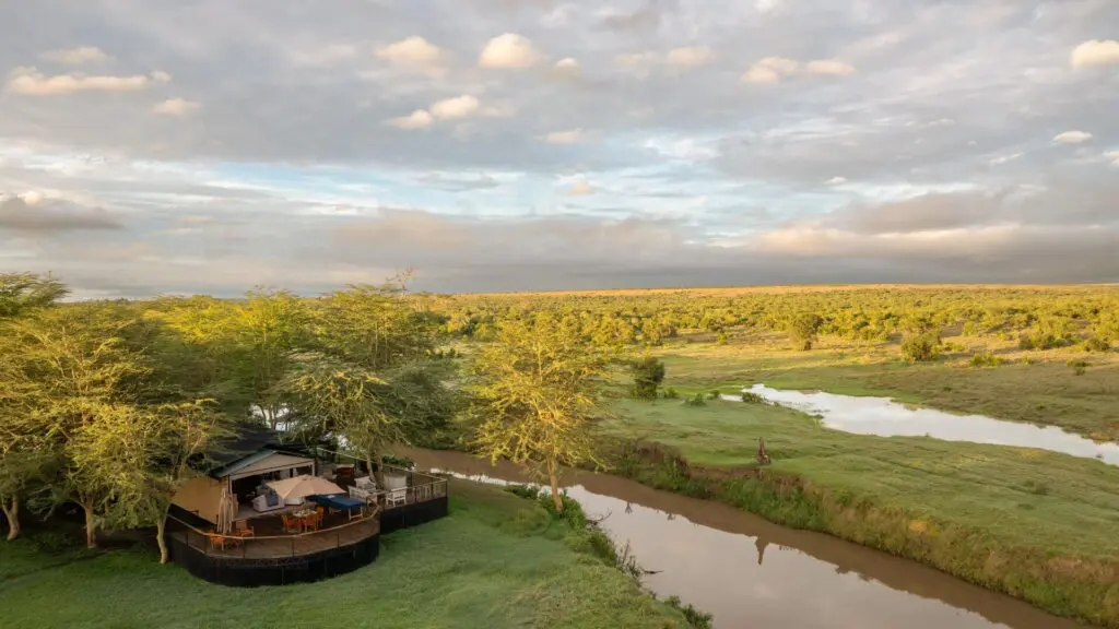 Une vue aérienne du camp Ol Pejeta Safari situé au bord d'une rivière sinueuse, avec des terrasses en bois nichées parmi les arbres et une vaste savane verte s'étendant à l'horizon sous un ciel doux et nuageux.