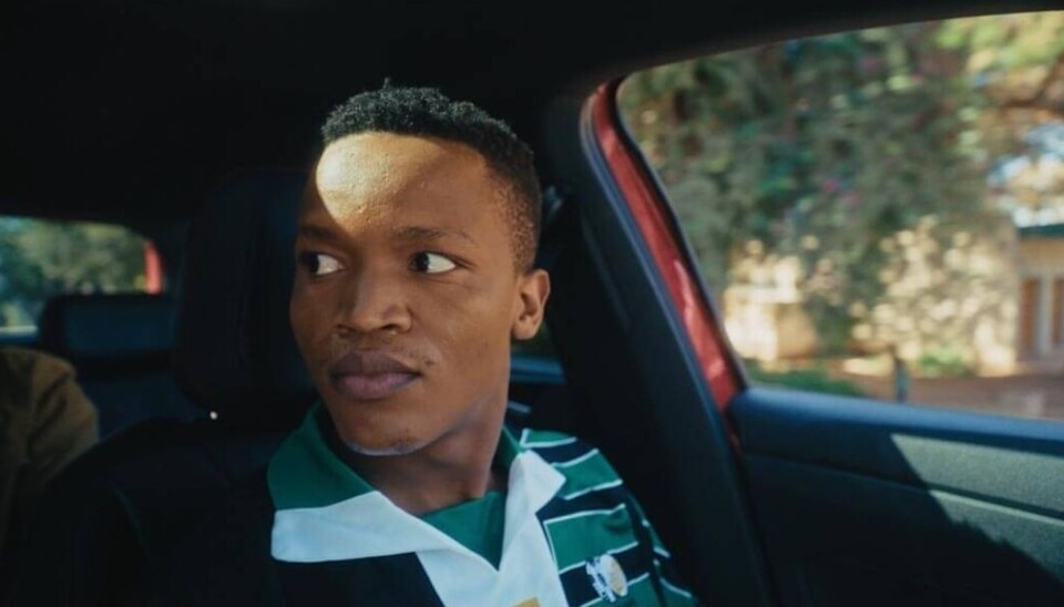 Image promotionnelle de « Ubani Lore » montrant un homme vêtu d'une chemise à rayures vertes et noires, assis dans une voiture et regardant de côté.
