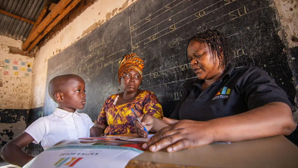 Un employé de Kafika House vêtu d'un polo de marque est assis avec un jeune enfant et une femme à un bureau dans une salle de classe rurale, examinant du matériel pédagogique devant un tableau sur lequel sont écrites des leçons d'anglais, Afrique de l'Est.