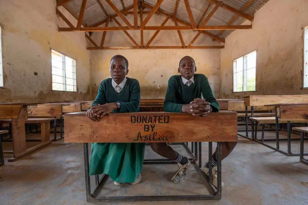 Deux étudiants en uniforme scolaire vert sont assis côte à côte devant un bureau en bois estampillé "Don d'Asilia" dans une salle de classe rurale remplie de pupitres assortis, Afrique de l'Est.