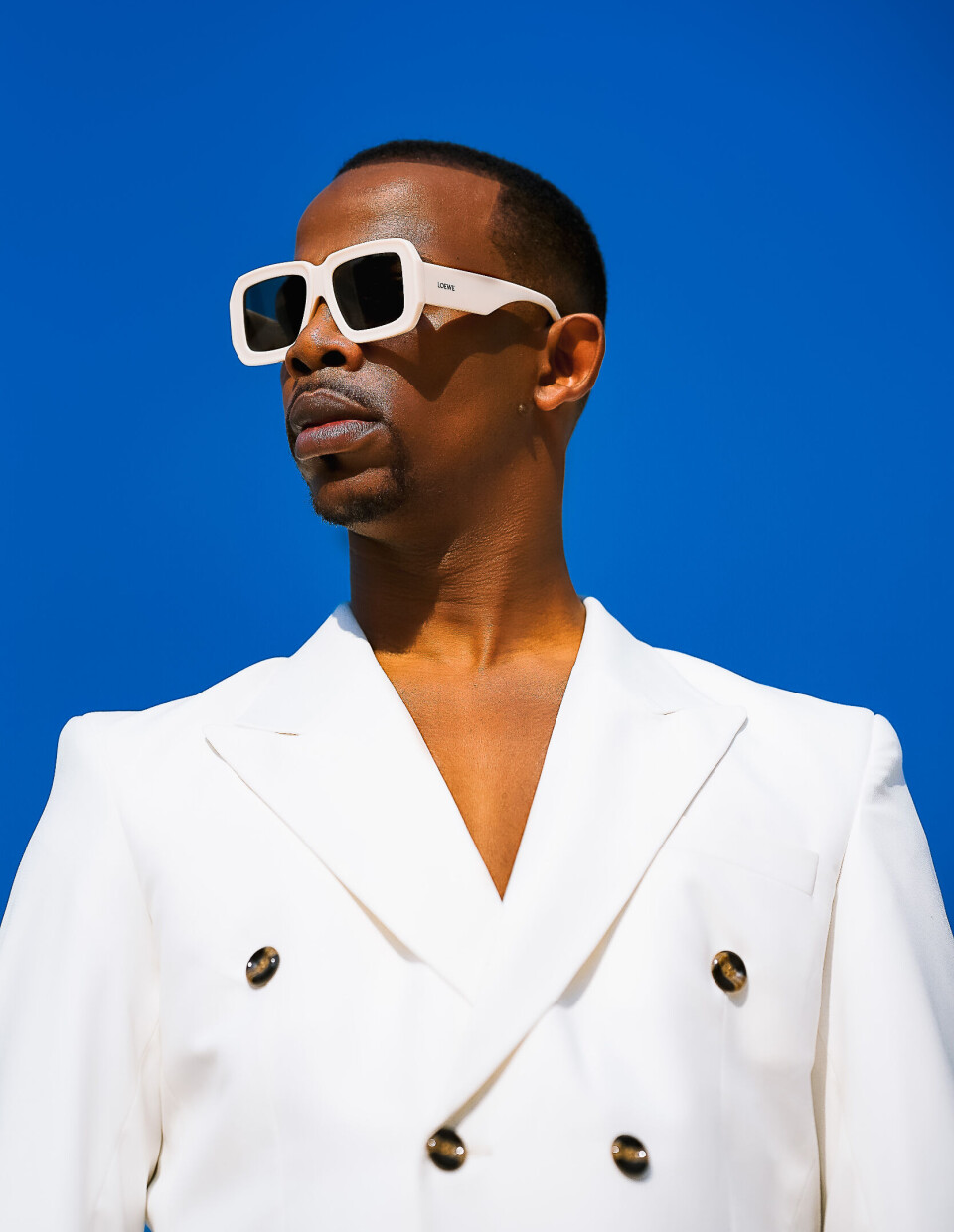 Une photo promotionnelle de Zakes Bantwini sur fond bleu ciel. Il porte des lunettes de soleil sombres avec une monture blanche et un blazer blanc.