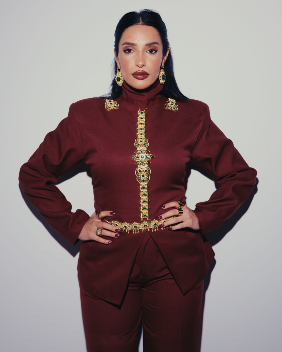 Inez porte un costume marocain rouge avec de gros bijoux dorés et regarde directement la caméra.