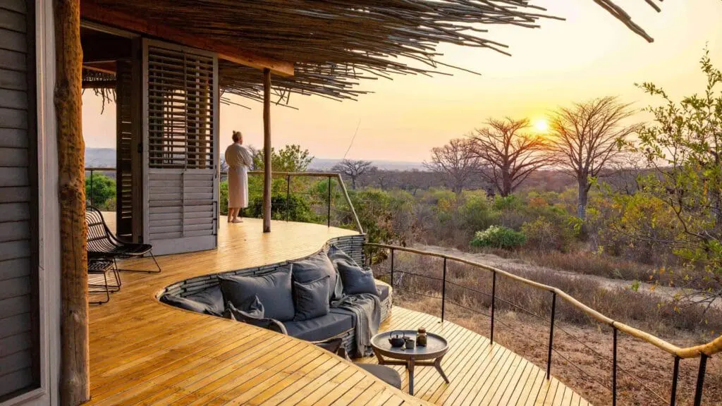 Un invité se tient sur une terrasse privée en bois au coucher du soleil, surplombant une forêt ouverte et des baobabs, avec des sièges intégrés et des tissus d'ameublement faisant face à la vue.