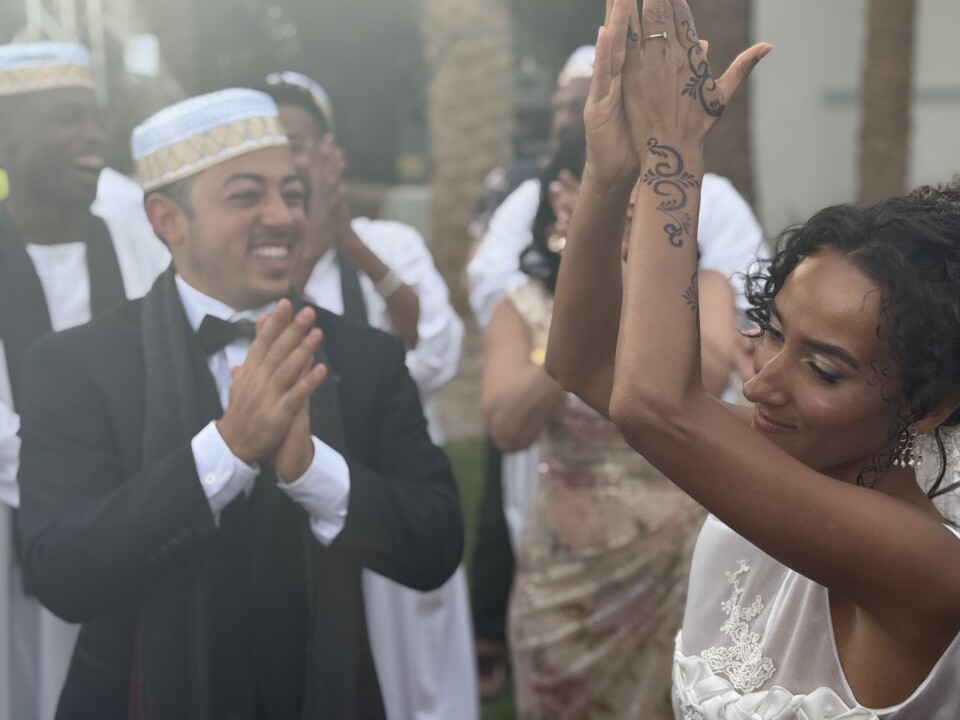 Un homme vêtu d’un costume noir et d’un chapeau soudanais et une femme vêtue d’une robe de mariée blanche sourient et dansent dans un jardin.
