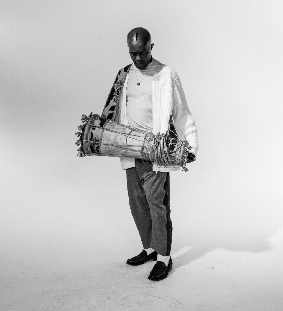 Une image de Sola Akingbola tenant un tambour. Il se tient sur un fond blanc.