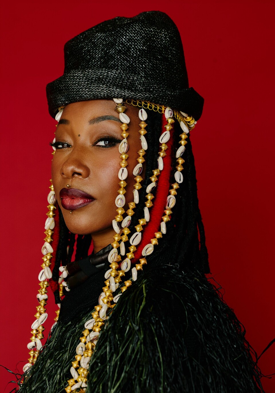 Portrait de Fatoumata Diawara au chapeau noir aux tresses ornées de coquillages sur fond rouge.