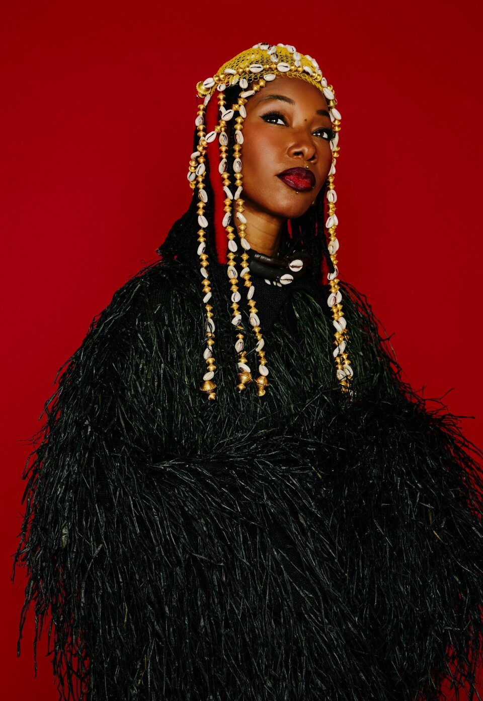 Portrait en studio de Fatoumata Diawara dans une tenue texturée noire et une coiffe perlée sur fond rouge.