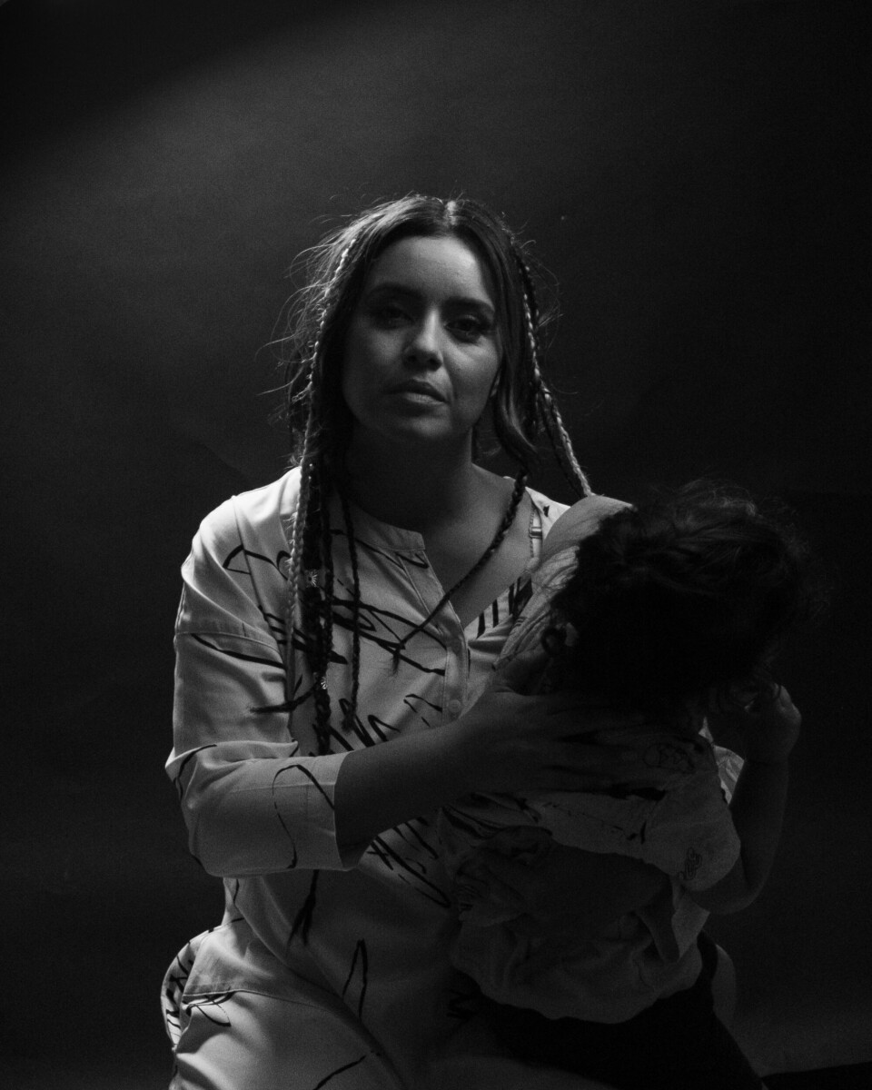 Image en noir et blanc de Neta tenant un enfant dans ses bras tout en regardant directement la caméra avec une expression faciale sérieuse.